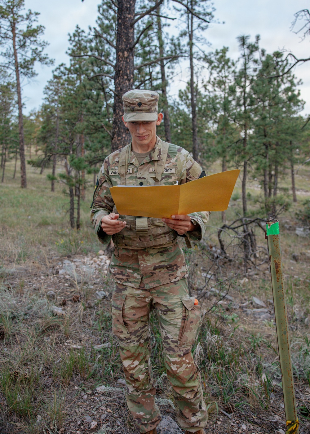 A U.S. Army soldier in OCP (Operational Camouflage Pattern) uniform is standing outdoors in a wooded, rocky terrain while reading documents from a manila/yellow folder. The soldier's name tape partially reads 'VAN...RT' and the branch tape reads 'U.S. ARMY'. A green-topped stake or marker post is visible to the soldier's right, suggesting a land navigation or orienteering exercise.