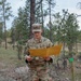 A U.S. Army soldier in OCP (Operational Camouflage Pattern) uniform is standing outdoors in a wooded, rocky terrain while reading documents from a manila/yellow folder. The soldier's name tape partially reads 'VAN...RT' and the branch tape reads 'U.S. ARMY'. A green-topped stake or marker post is visible to the soldier's right, suggesting a land navigation or orienteering exercise.