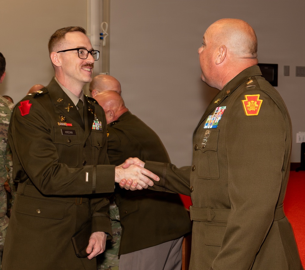 Two U.S. Army officers in Army Service Uniforms (green dress uniforms) are shaking hands at what appears to be a formal military ceremony or event. The officer on the left, identified by his name tape as 'KRZAN,' is smiling and wearing glasses, while the officer on the right displays a Pennsylvania National Guard shoulder patch. Additional military personnel are visible in the background.