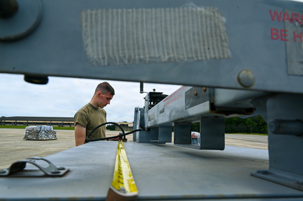 A military service member in an olive drab t-shirt is working on or inspecting ground support equipment on an airfield tarmac. The photo is taken from a low angle through the frame of the equipment, creating a framed perspective. The setting appears to be a military air base with hangar structures and tree line visible in the background.