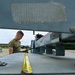 A military service member in an olive drab t-shirt is working on or inspecting ground support equipment on an airfield tarmac. The photo is taken from a low angle through the frame of the equipment, creating a framed perspective. The setting appears to be a military air base with hangar structures and tree line visible in the background.