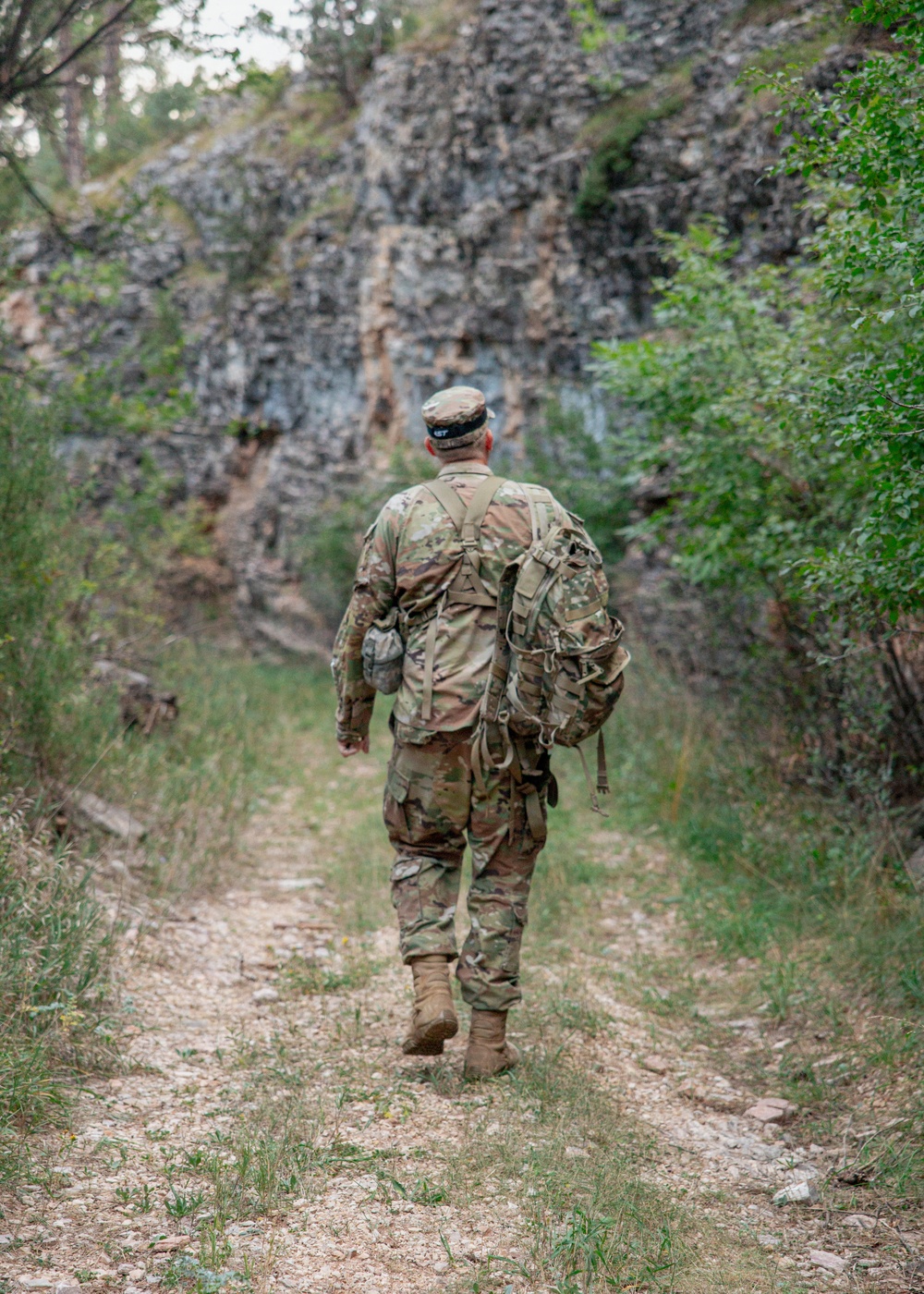 A soldier in full camouflage uniform and tactical gear walks away from the camera along a narrow dirt trail. The path leads toward a rocky cliff face flanked by green vegetation and shrubs. The image captures a rucksack march or field training exercise in a rugged outdoor environment.