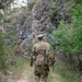 A soldier in full camouflage uniform and tactical gear walks away from the camera along a narrow dirt trail. The path leads toward a rocky cliff face flanked by green vegetation and shrubs. The image captures a rucksack march or field training exercise in a rugged outdoor environment.