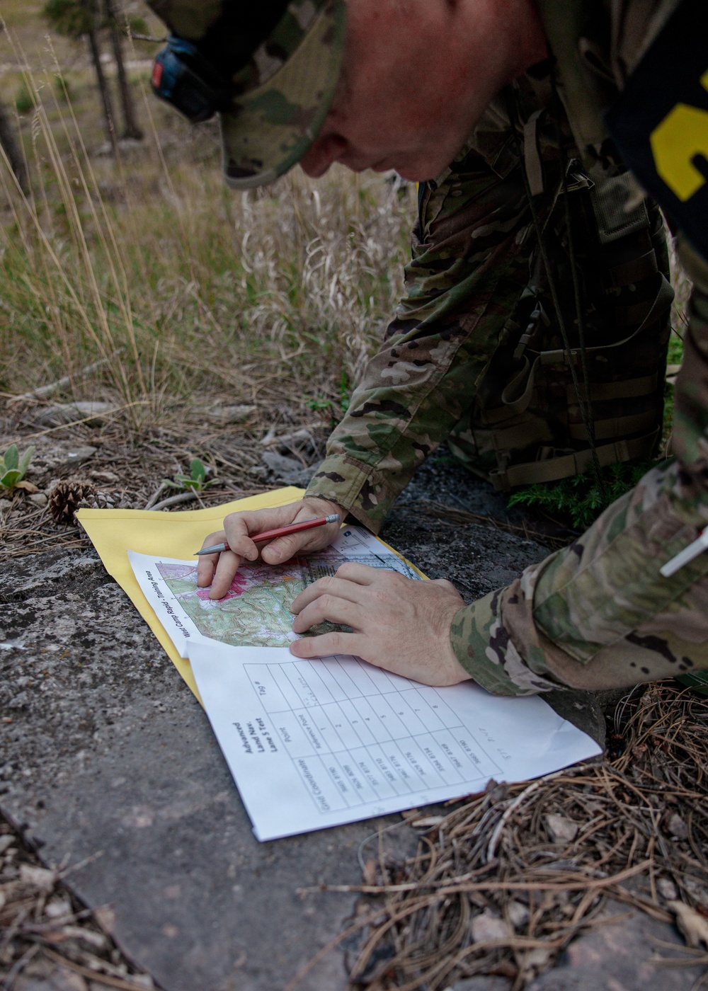 A soldier in MultiCam/OCP camouflage uniform is kneeling on a rock surface, marking or plotting points on a topographic map. The documents are placed on a yellow folder/envelope, and the soldier is using a pencil to record information on an accompanying data sheet. This appears to be a land navigation training exercise based on the visible documents.