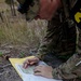 A soldier in MultiCam/OCP camouflage uniform is kneeling on a rock surface, marking or plotting points on a topographic map. The documents are placed on a yellow folder/envelope, and the soldier is using a pencil to record information on an accompanying data sheet. This appears to be a land navigation training exercise based on the visible documents.