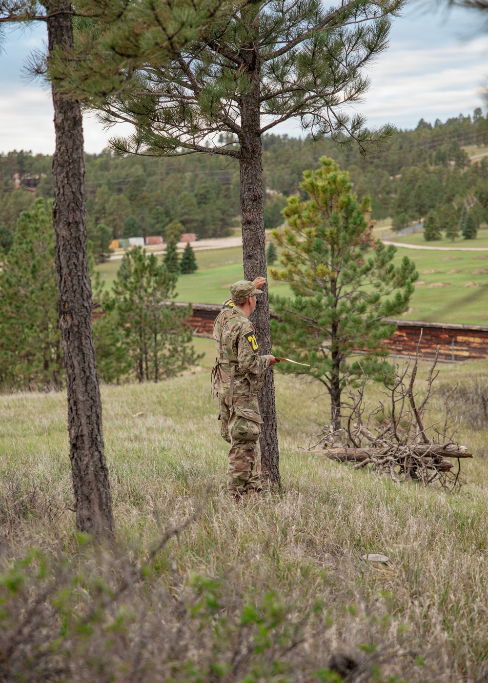 A person in military camouflage uniform with a yellow number identifier on their back stands near a pine tree on a grassy hillside, appearing to consult a document or map. The setting appears to be a military training exercise conducted in a rural, forested landscape with open meadows and rustic wooden structures visible in the background. The terrain and vegetation suggest a high-altitude or mountainous western U.S. environment.