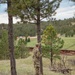 A person in military camouflage uniform with a yellow number identifier on their back stands near a pine tree on a grassy hillside, appearing to consult a document or map. The setting appears to be a military training exercise conducted in a rural, forested landscape with open meadows and rustic wooden structures visible in the background. The terrain and vegetation suggest a high-altitude or mountainous western U.S. environment.