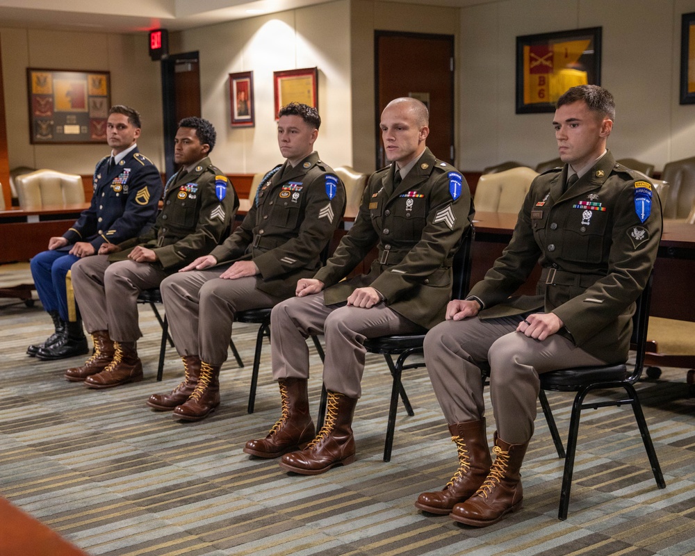Five U.S. Army soldiers are seated in a row on black chairs in what appears to be a formal military conference or ceremony room. Four soldiers wear olive drab Class A dress uniforms with various patches, medals, and brown jump boots, while one soldier on the far left wears the blue Army Service Uniform. The setting includes framed military unit insignia and plaques on the walls, suggesting an official awards or recognition ceremony.