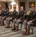 Five U.S. Army soldiers are seated in a row on black chairs in what appears to be a formal military conference or ceremony room. Four soldiers wear olive drab Class A dress uniforms with various patches, medals, and brown jump boots, while one soldier on the far left wears the blue Army Service Uniform. The setting includes framed military unit insignia and plaques on the walls, suggesting an official awards or recognition ceremony.