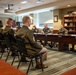 A formal military briefing or meeting is taking place in what appears to be a senior officer's conference room. A group of lower-ranking soldiers seated in chairs on the left side face a panel of higher-ranking officers seated at a large conference table on the right. The setting suggests an official Army meeting, possibly a mentorship session, board review, or command briefing.
