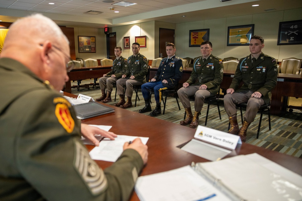 A senior non-commissioned officer, identified by a name placard as 'SGM Vern Daley', conducts what appears to be a formal board or evaluation panel with five seated soldiers in a military conference room. The soldiers are dressed in various U.S. Army uniforms including dress blues and Army Service Uniforms (ASU/greens), suggesting a promotion or competition board setting. The room is decorated with military unit guidons and framed documents on the walls.