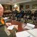 A senior non-commissioned officer, identified by a name placard as 'SGM Vern Daley', conducts what appears to be a formal board or evaluation panel with five seated soldiers in a military conference room. The soldiers are dressed in various U.S. Army uniforms including dress blues and Army Service Uniforms (ASU/greens), suggesting a promotion or competition board setting. The room is decorated with military unit guidons and framed documents on the walls.