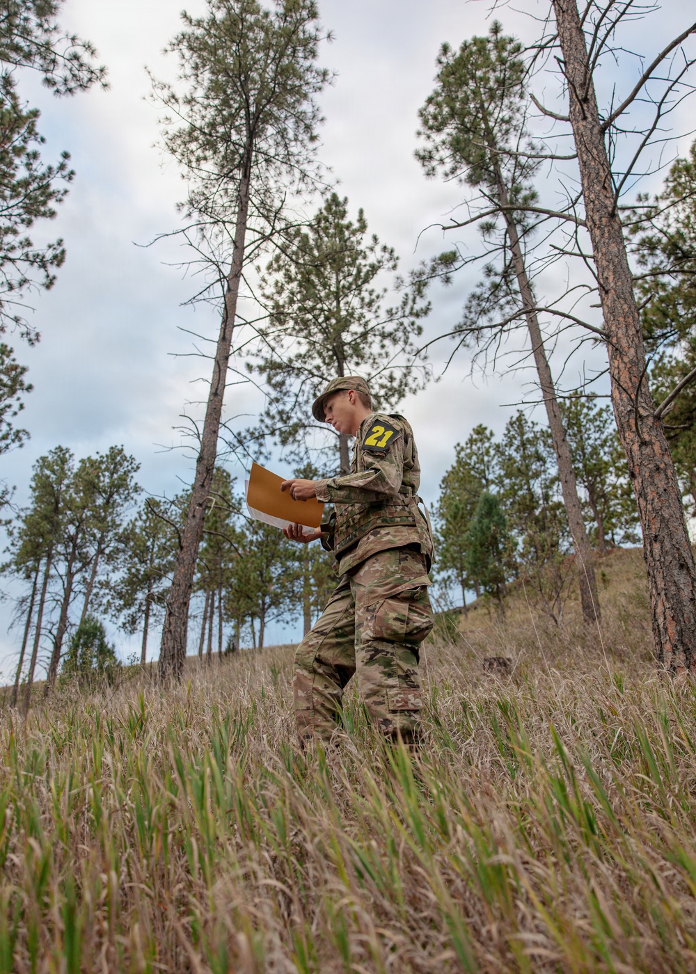 A soldier or military cadet wearing multicam/OCP camouflage uniform and a patrol cap is standing in a grassy, wooded hillside area reviewing documents or a map. The individual has a yellow number '21' patch visible on their left sleeve, suggesting participation in a timed exercise or competition. The setting appears to be a land navigation or orienteering training exercise in a pine forest environment.
