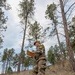 A soldier or military cadet wearing multicam/OCP camouflage uniform and a patrol cap is standing in a grassy, wooded hillside area reviewing documents or a map. The individual has a yellow number '21' patch visible on their left sleeve, suggesting participation in a timed exercise or competition. The setting appears to be a land navigation or orienteering training exercise in a pine forest environment.