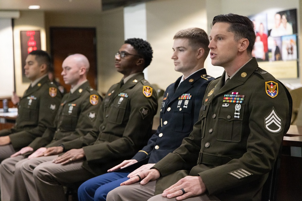 Five U.S. Army soldiers in dress green uniforms are seated in a row, appearing to be at a formal ceremony or official proceeding indoors. The soldiers display various ranks and ribbons, with the foreground soldier wearing Staff Sergeant chevrons and multiple service ribbons. They appear attentive, possibly listening to or participating in an official event.