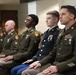 Five U.S. Army soldiers in dress green uniforms are seated in a row, appearing to be at a formal ceremony or official proceeding indoors. The soldiers display various ranks and ribbons, with the foreground soldier wearing Staff Sergeant chevrons and multiple service ribbons. They appear attentive, possibly listening to or participating in an official event.