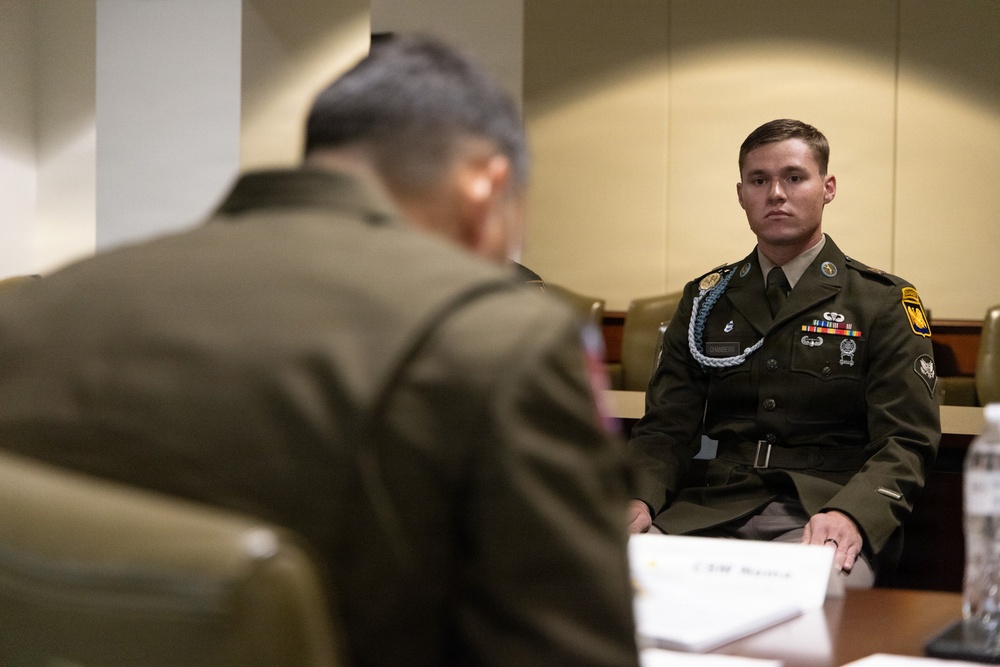 Two military personnel in olive green dress uniforms are seated at a conference table in what appears to be a formal meeting room. The soldier in focus on the right is wearing multiple decorations including ribbons, badges, a blue shoulder cord (aiguillette), and unit patches on his uniform. A name placard is partially visible on the table in the foreground.