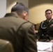 Two military personnel in olive green dress uniforms are seated at a conference table in what appears to be a formal meeting room. The soldier in focus on the right is wearing multiple decorations including ribbons, badges, a blue shoulder cord (aiguillette), and unit patches on his uniform. A name placard is partially visible on the table in the foreground.