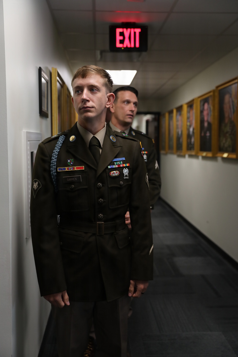 Two U.S. Army soldiers in Army Service Uniform (Class A dress uniform) stand in a hallway lined with framed portraits. The foreground soldier, identified by nameplate as 'GARREN,' wears multiple service ribbons, badges, and a blue cord (infantry). An illuminated EXIT sign is visible overhead.