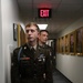 Two U.S. Army soldiers in Army Service Uniform (Class A dress uniform) stand in a hallway lined with framed portraits. The foreground soldier, identified by nameplate as 'GARREN,' wears multiple service ribbons, badges, and a blue cord (infantry). An illuminated EXIT sign is visible overhead.