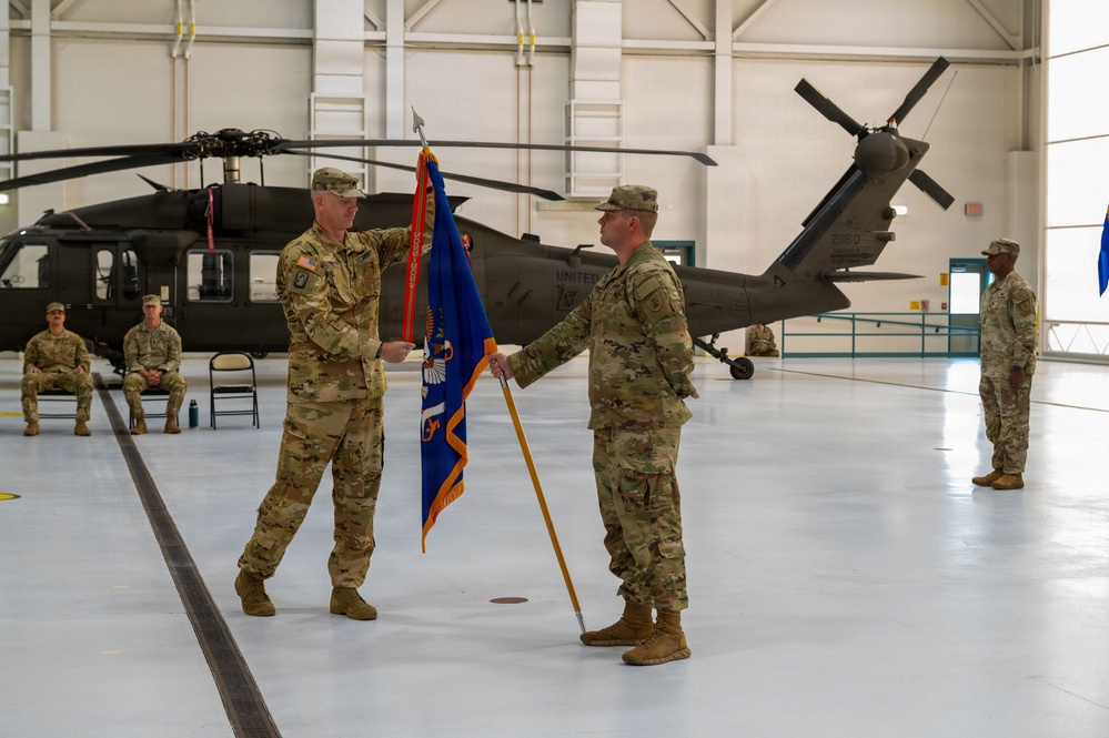 A military change of command ceremony is taking place inside a helicopter hangar. Two soldiers in U.S. Army OCP (Operational Camouflage Pattern) uniforms are exchanging a blue unit guidon flag, which is the central symbolic act of a command transfer. Black UH-60 Black Hawk helicopters are visible in the background, along with several seated and standing soldiers observing the ceremony.