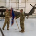 A military change of command ceremony is taking place inside a helicopter hangar. Two soldiers in U.S. Army OCP (Operational Camouflage Pattern) uniforms are exchanging a blue unit guidon flag, which is the central symbolic act of a command transfer. Black UH-60 Black Hawk helicopters are visible in the background, along with several seated and standing soldiers observing the ceremony.