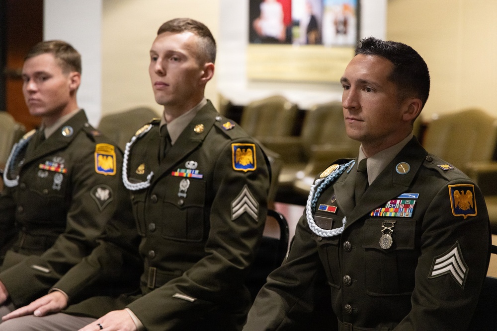 Three U.S. Army soldiers in dress green uniforms are seated in what appears to be a formal ceremony or official event. All three wear the same yellow-and-black shoulder patch (consistent with the 10th Mountain Division or similar unit) and display various medals, ribbons, and braided aiguillettes. The soldiers appear attentive, facing forward in a formal setting.
