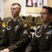 Three U.S. Army soldiers in dress green uniforms are seated in what appears to be a formal ceremony or official event. All three wear the same yellow-and-black shoulder patch (consistent with the 10th Mountain Division or similar unit) and display various medals, ribbons, and braided aiguillettes. The soldiers appear attentive, facing forward in a formal setting.