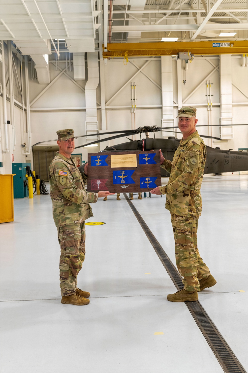 Two U.S. Army soldiers in OCP (Operational Camouflage Pattern) uniforms are standing in a military aviation hangar, jointly holding a wooden commemorative plaque mounted with multiple unit guidon pennants. The plaque displays several blue and red guidons with unit designations including '112' and '285', along with a central brass inscription plate. A UH-60 Black Hawk helicopter marked 'UNITED STATES' is visible in the background.