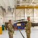 Two U.S. Army soldiers in OCP (Operational Camouflage Pattern) uniforms are standing in a military aviation hangar, jointly holding a wooden commemorative plaque mounted with multiple unit guidon pennants. The plaque displays several blue and red guidons with unit designations including '112' and '285', along with a central brass inscription plate. A UH-60 Black Hawk helicopter marked 'UNITED STATES' is visible in the background.
