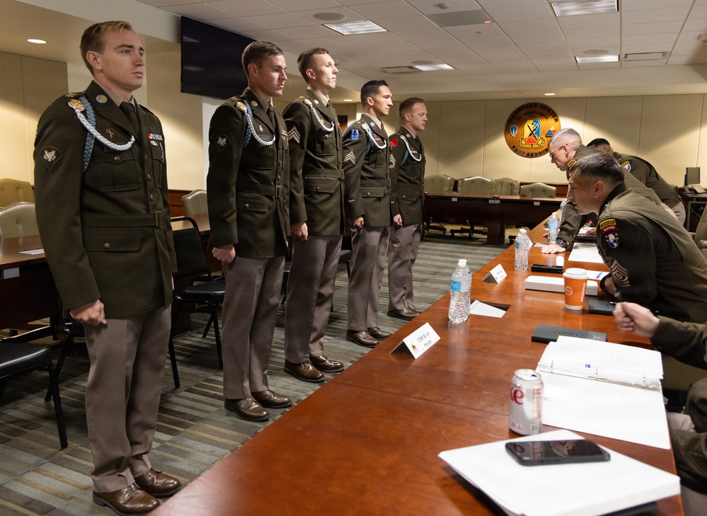 A formal military board or evaluation proceeding is taking place in a conference room, with four U.S. Army soldiers in dress green uniforms standing at attention before a panel of senior officers seated at a long wooden table. The soldiers are wearing aiguillettes (ceremonial cords) and various rank insignia, suggesting this may be a promotion board, competition board, or formal inspection. A unit crest/insignia is visible on the wall in the background.