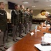 A formal military board or evaluation proceeding is taking place in a conference room, with four U.S. Army soldiers in dress green uniforms standing at attention before a panel of senior officers seated at a long wooden table. The soldiers are wearing aiguillettes (ceremonial cords) and various rank insignia, suggesting this may be a promotion board, competition board, or formal inspection. A unit crest/insignia is visible on the wall in the background.