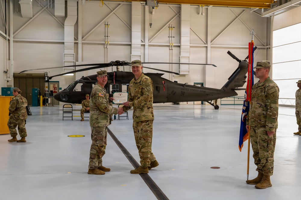 A military ceremony is taking place inside a United States Army helicopter hangar. Two soldiers in OCP (Operational Camouflage Pattern) uniforms are shaking hands while exchanging what appears to be a certificate or award, with a UH-60 Black Hawk helicopter visible in the background. Additional soldiers are present in formation, and a unit guidon/flag is being held by a soldier on the right.