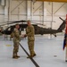A military ceremony is taking place inside a United States Army helicopter hangar. Two soldiers in OCP (Operational Camouflage Pattern) uniforms are shaking hands while exchanging what appears to be a certificate or award, with a UH-60 Black Hawk helicopter visible in the background. Additional soldiers are present in formation, and a unit guidon/flag is being held by a soldier on the right.
