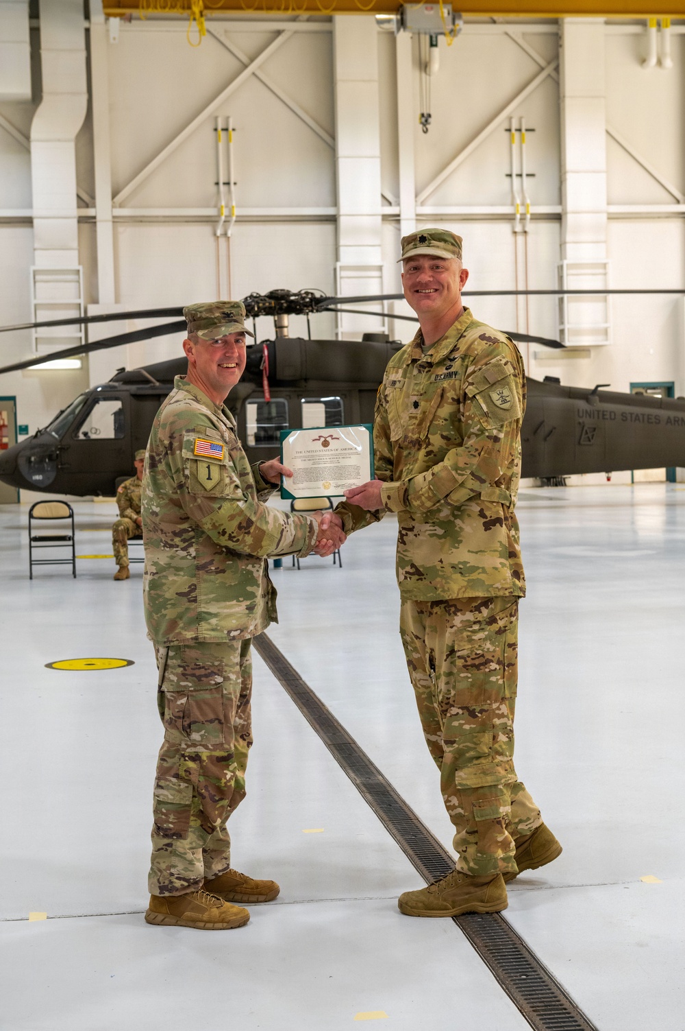 Two U.S. Army soldiers in OCP (Operational Camouflage Pattern) uniforms are shaking hands while jointly holding a certificate or award document inside a military aviation hangar. A UH-60 Black Hawk helicopter marked 'United States A[rmy]' is visible in the background. This appears to be a formal military awards or promotion ceremony.