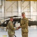 Two U.S. Army soldiers in OCP (Operational Camouflage Pattern) uniforms are shaking hands while jointly holding a certificate or award document inside a military aviation hangar. A UH-60 Black Hawk helicopter marked 'United States A[rmy]' is visible in the background. This appears to be a formal military awards or promotion ceremony.