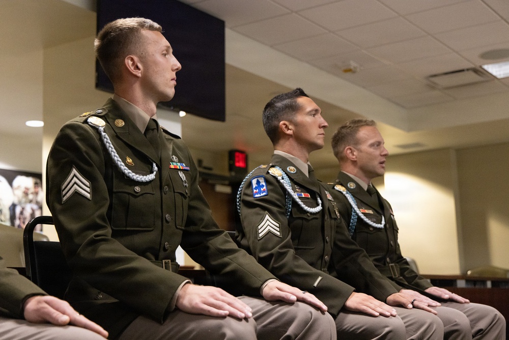 Three U.S. Army soldiers in dress green uniforms are seated in a row at what appears to be a formal military ceremony or award presentation. All three wear blue fourragères (shoulder cords), service ribbons, and rank insignia consistent with the rank of Sergeant. They are seated at attention, facing forward in an indoor military facility.