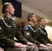Three U.S. Army soldiers in dress green uniforms are seated in a row at what appears to be a formal military ceremony or award presentation. All three wear blue fourragères (shoulder cords), service ribbons, and rank insignia consistent with the rank of Sergeant. They are seated at attention, facing forward in an indoor military facility.