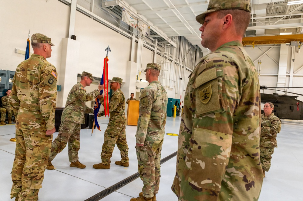 A military change of command or activation ceremony is taking place inside a large hangar facility. Soldiers in U.S. Army OCP (Operational Camouflage Pattern) uniforms are participating in a guidon transfer, which is the central ritual of such ceremonies. A military helicopter is visible in the background, confirming the aviation unit setting.