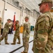 A military change of command or activation ceremony is taking place inside a large hangar facility. Soldiers in U.S. Army OCP (Operational Camouflage Pattern) uniforms are participating in a guidon transfer, which is the central ritual of such ceremonies. A military helicopter is visible in the background, confirming the aviation unit setting.