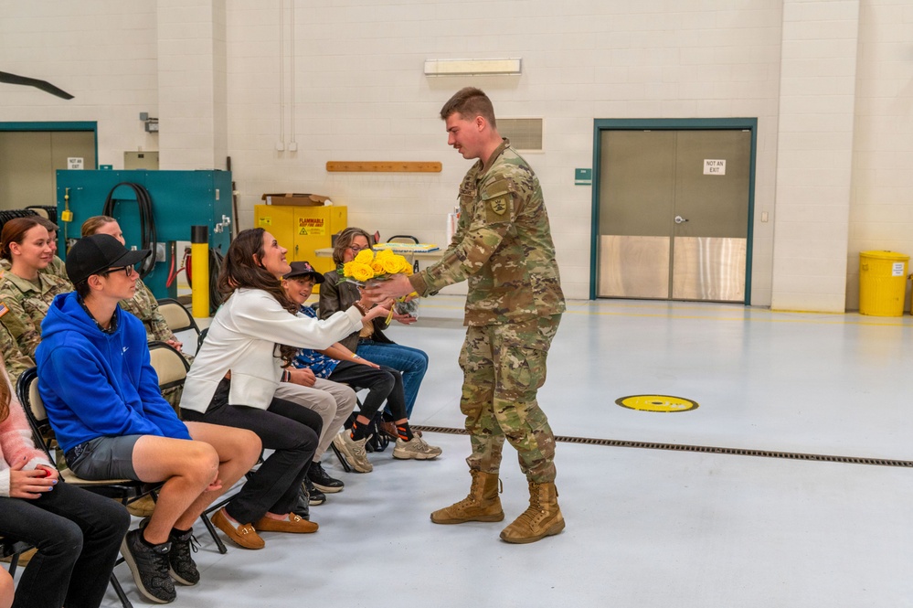 A soldier in US Army OCP (Operational Camouflage Pattern) uniform is presenting a bouquet of yellow roses to a seated woman in a white jacket during what appears to be a military ceremony or event. The setting is an indoor military facility or hangar with other attendees, including civilians and uniformed personnel, seated in folding chairs. The atmosphere appears warm and celebratory.