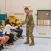 A soldier in US Army OCP (Operational Camouflage Pattern) uniform is presenting a bouquet of yellow roses to a seated woman in a white jacket during what appears to be a military ceremony or event. The setting is an indoor military facility or hangar with other attendees, including civilians and uniformed personnel, seated in folding chairs. The atmosphere appears warm and celebratory.