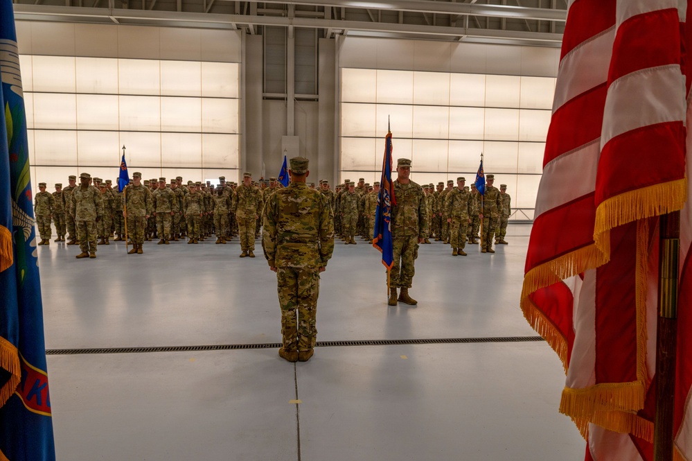 A formal military ceremony is taking place inside a large aircraft hangar. Uniformed personnel in OCP (Operational Camouflage Pattern) uniforms are arranged in formation facing a central figure, with flag bearers holding unit and national colors. The setting and arrangement suggest a change of command, activation, or similar official military ceremony.
