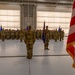 A formal military ceremony is taking place inside a large aircraft hangar. Uniformed personnel in OCP (Operational Camouflage Pattern) uniforms are arranged in formation facing a central figure, with flag bearers holding unit and national colors. The setting and arrangement suggest a change of command, activation, or similar official military ceremony.