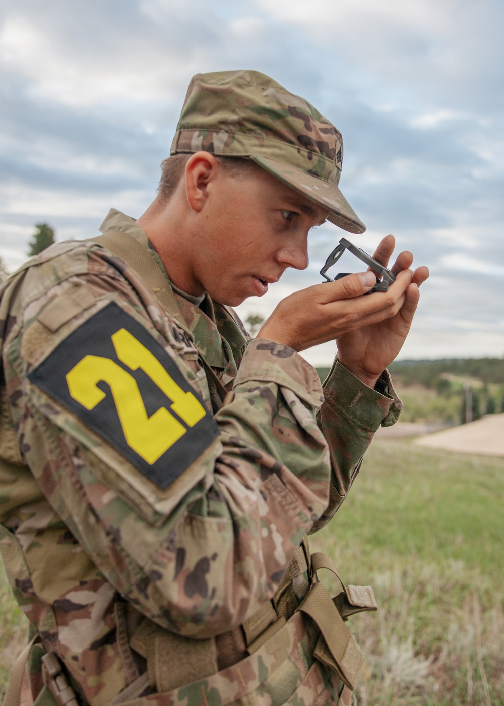 A young soldier in US Army OCP (Operational Camouflage Pattern) uniform is using a lensatic compass, holding it up to eye level to take a bearing or azimuth reading. The soldier wears a black and yellow '21' competition number patch on the left sleeve, suggesting participation in a land navigation competition or military skills event. The setting appears to be an outdoor training area with open terrain and overcast skies.