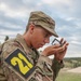 A young soldier in US Army OCP (Operational Camouflage Pattern) uniform is using a lensatic compass, holding it up to eye level to take a bearing or azimuth reading. The soldier wears a black and yellow '21' competition number patch on the left sleeve, suggesting participation in a land navigation competition or military skills event. The setting appears to be an outdoor training area with open terrain and overcast skies.