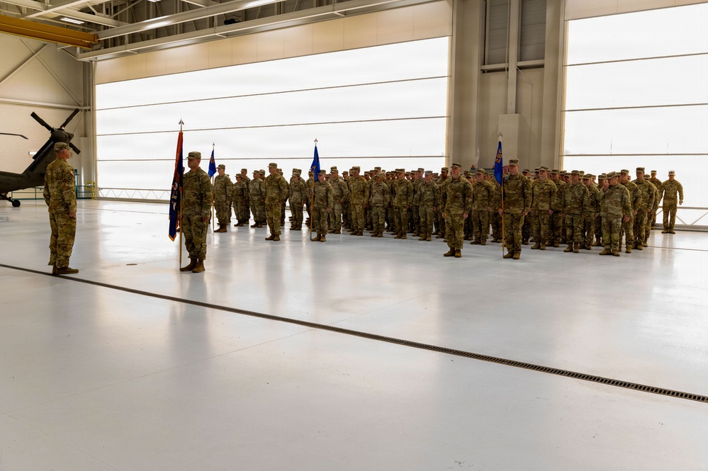 A military formation ceremony is taking place inside a large aircraft hangar. Numerous soldiers in camouflage uniforms are standing at attention in organized ranks, with flag bearers holding unit guidons. A single officer stands facing the formation, suggesting a formal military ceremony such as a change of command, activation, or unit review.