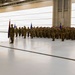 A military formation ceremony is taking place inside a large aircraft hangar. Numerous soldiers in camouflage uniforms are standing at attention in organized ranks, with flag bearers holding unit guidons. A single officer stands facing the formation, suggesting a formal military ceremony such as a change of command, activation, or unit review.