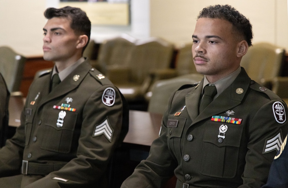 Two young male soldiers in U.S. Army dress green uniforms are seated in what appears to be an official ceremony or formal military setting. Both wear multiple service ribbons, medals, rank insignia (Sergeant chevrons), and unit patches on their left shoulders. They are seated attentively, facing forward.