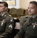 Two young male soldiers in U.S. Army dress green uniforms are seated in what appears to be an official ceremony or formal military setting. Both wear multiple service ribbons, medals, rank insignia (Sergeant chevrons), and unit patches on their left shoulders. They are seated attentively, facing forward.